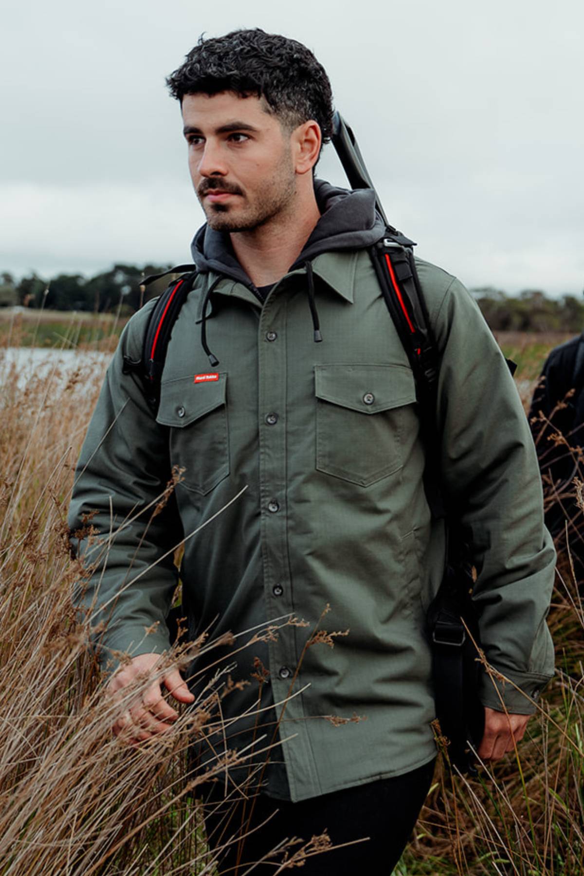 A person with dark hair and a short beard is walking through tall grass. They are wearing the Hard Yakka Quilted Solid Shacket made from durable gray ripstop fabric, a black hoodie, and carrying a black-and-red backpack. The background includes trees and a cloudy sky.