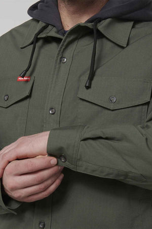 A person wearing the Hard Yakka Quilted Solid Shacket in olive-green, with black drawstrings visible from the hooded sweatshirt underneath. The shacket, made from durable ripstop fabric, features buttoned chest pockets and the person is fastening the cuff of their left sleeve. A small red label is sewn on one of the pockets.