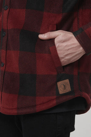 A person with a hand in their pocket models the Hard Yakka Sherpa Bonded Fleece Jacket, featuring a red and black checkered pattern. The jacket is designed with black buttons, adjustable cuffs, sherpa lining, and a brown stitched label near the hem. The person completes the look with black pants.