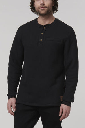 A man models the Hard Yakka Sherpa Henley, a regular fit black long-sleeve shirt with a buttoned collar. It has a small patch near the hem, and he stands against a neutral backdrop with his hands relaxed at his sides.