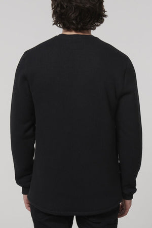 A person with dark wavy hair wears a long-sleeve black Hard Yakka Sherpa Henley, standing against a plain gray background.