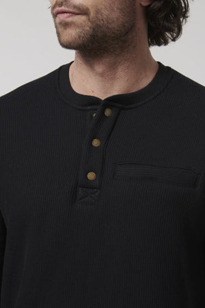 A man with medium-length hair wears a Hard Yakka Sherpa Henley—a regular fit, black shirt featuring three visible brass buttons and a small chest pocket. The image highlights his upper torso and part of his face with a neutral expression.