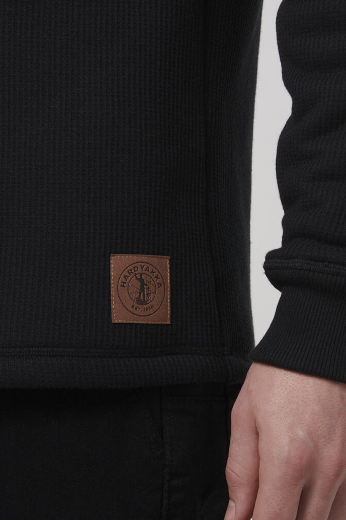 A person is wearing the Hard Yakka Sherpa Henley, a black ribbed sweater with sherpa lining. A small brown patch featuring a circular logo and "HARD YAKKA EST 1934" is near the hem. Their slightly bent arm highlights the garment's cozy texture.