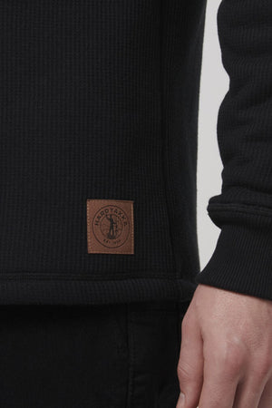 A person is wearing the Hard Yakka Sherpa Henley, a black ribbed sweater with sherpa lining. A small brown patch featuring a circular logo and "HARD YAKKA EST 1934" is near the hem. Their slightly bent arm highlights the garment's cozy texture.