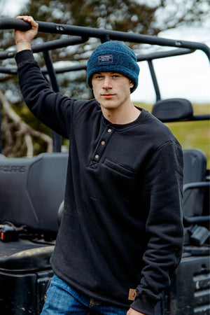 Wearing a blue knit beanie and a Hard Yakka Sherpa Henley, a person leans against an off-road vehicle in an outdoor setting with grassy terrain and trees.
