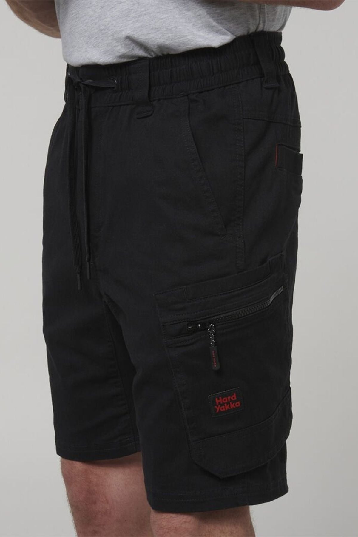 A person wears the Hard Yakka Tough Maxx Mid Short, featuring UPF 50+, multiple pockets including a zippered one with a red Hard Yakka logo, plus an elastic waistband and a gray t-shirt tucked in.