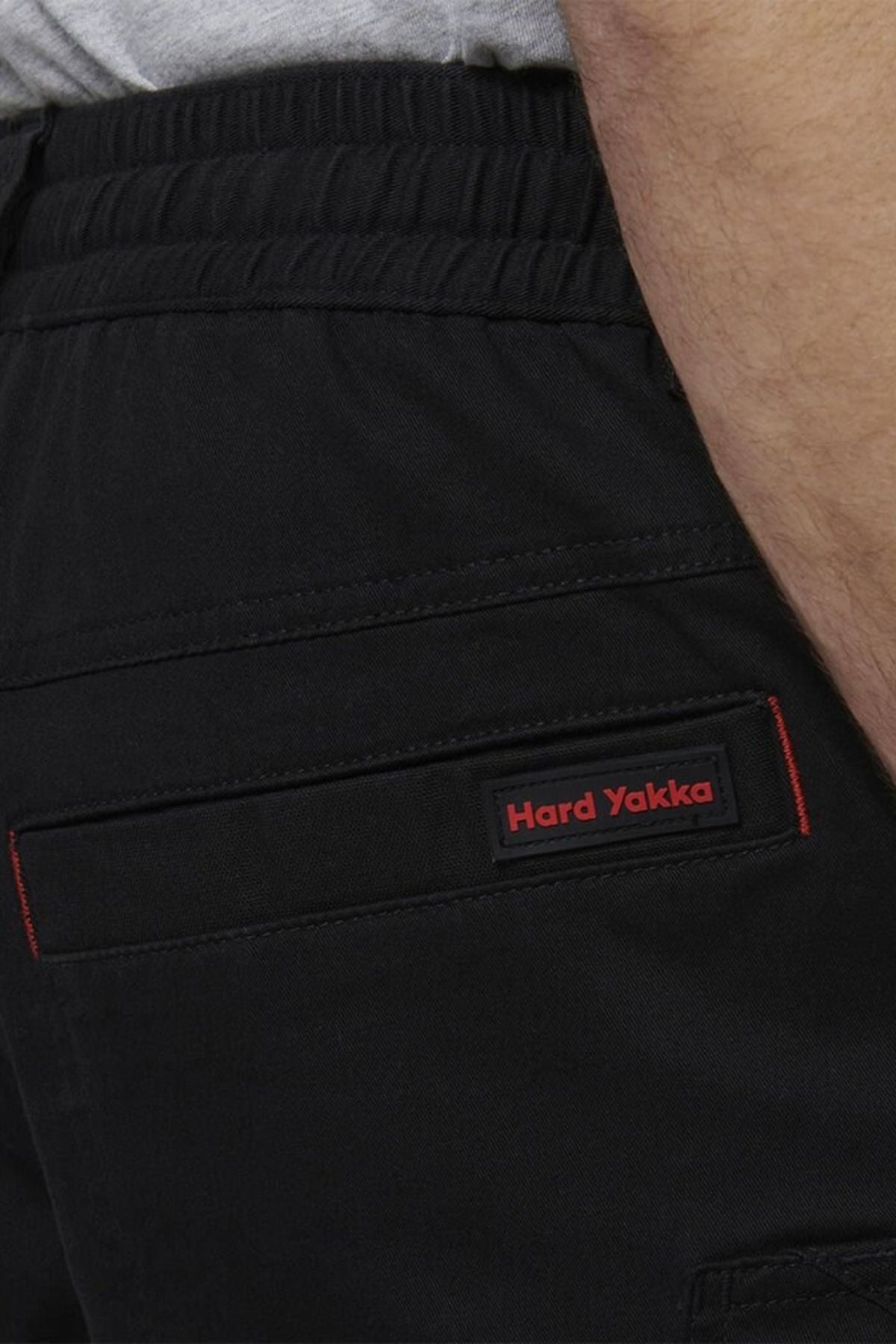Close-up of a person wearing Hard Yakka Tough Maxx Mid Short with an elastic waistband and a red and black label above the back pocket. Part of their arm and grey shirt are visible. Shorts offer UPF 50+ sun protection.
