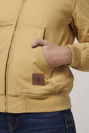 A person showcases the Hard Yakka Women's Bomber Jacket in tan, featuring a zipper and leather patch on the pocket. Their hand casually rests inside, paired with blue jeans, and the elegant fit is highlighted against a light-colored background.
