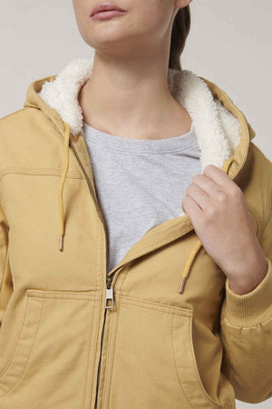A woman in a partially unzipped beige Hard Yakka Women's Bomber Jacket reveals a gray shirt underneath while adjusting the hood string. The background is a neutral light gray.