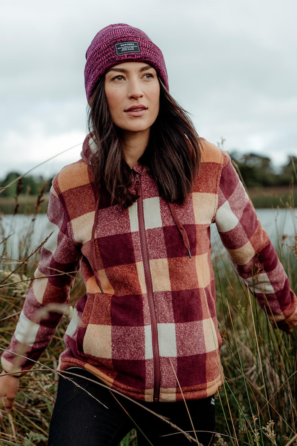 A woman in a Hard Yakka Womens Check Zoodie with a fleece body and thermal insulation, plus a purple beanie, stands outdoors in tall grass by a chilly lakeside under cloudy skies.