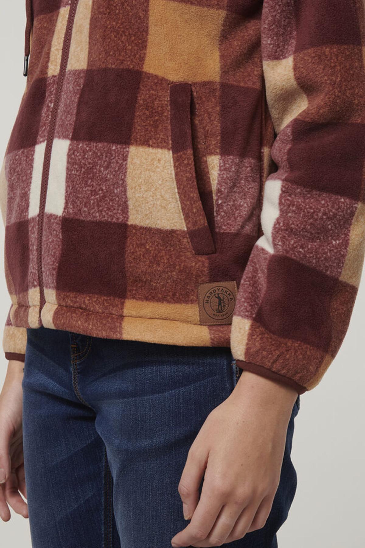 A person is shown wearing the Hard Yakka Womens Check Zoodie—a brown, tan, and white fleece jacket with a zipper—paired with blue jeans. The visible torso and arm highlight its cozy, thermally insulated fleece body.