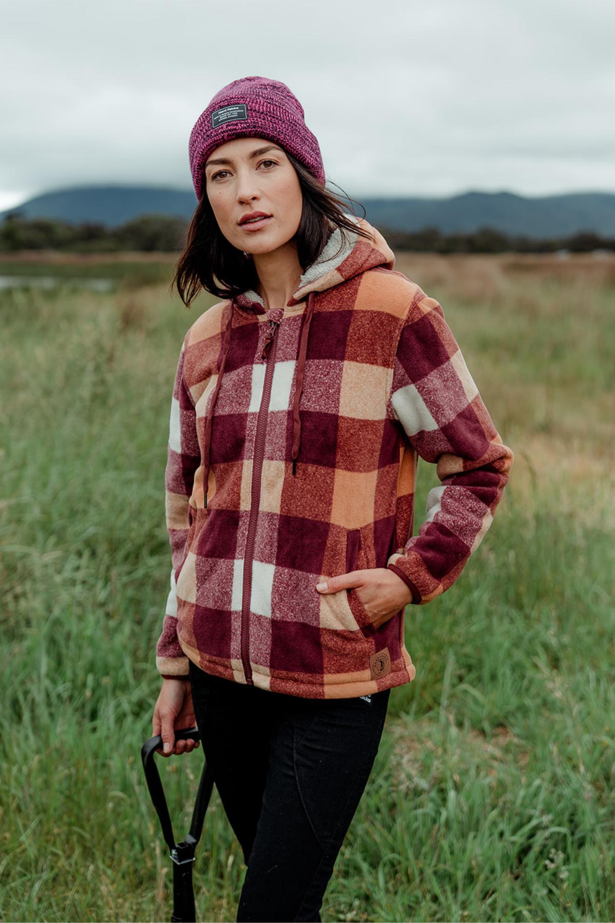 A woman outdoors in a grassy field holds a shovel while wearing a maroon and orange Hard Yakka Womens Check Zoodie with thermal insulation and a purple beanie, with mountains and cloudy skies in the background.