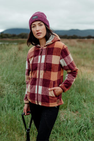 A woman outdoors in a grassy field holds a shovel while wearing a maroon and orange Hard Yakka Womens Check Zoodie with thermal insulation and a purple beanie, with mountains and cloudy skies in the background.