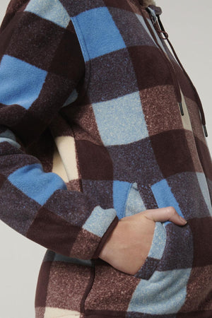 A close-up shows a person in the Hard Yakka Womens Check Zoodie, featuring a blue, brown, and cream check fleece with drawstrings and front pocket. The hoodie provides thermal insulation for warmth.