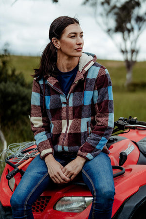 Wearing a Hard Yakka Womens Check Zoodie and blue jeans, a person sits on a red ATV outdoors. Green grass, trees, and a cloudy sky form the backdrop as they gaze to the side.