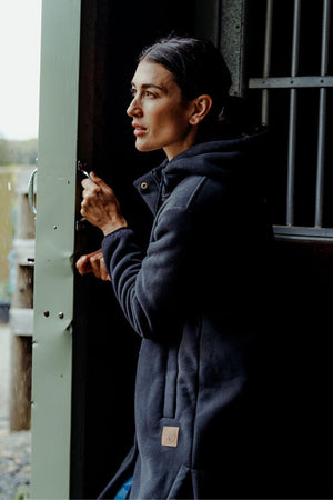 A woman in a dark Hard Yakka Womens Longline Zoodie with sherpa fleece stands indoors by an open door, thoughtfully looking outside while holding the handle as natural light streams in.