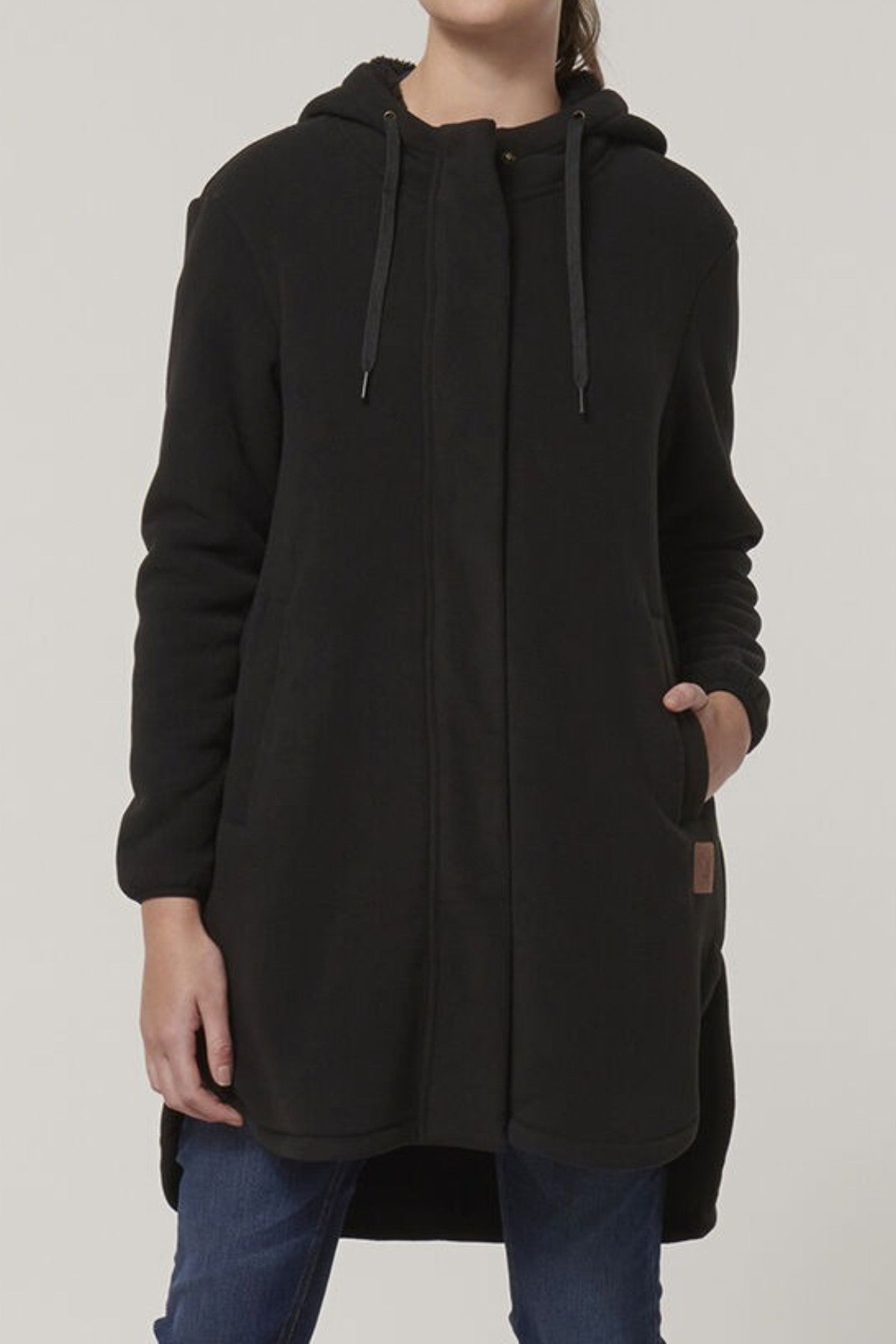 A person stands against a plain light background, wearing the Hard Yakka Womens Longline Zoodieβ a long, black sherpa-bonded fleece hooded coat with thermal insulationβ and blue jeans. Only the lower half of their face is visible; their left hand is in a pocket.
