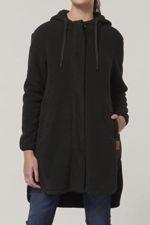A person stands against a plain light background, wearing the Hard Yakka Womens Longline Zoodieβ a long, black sherpa-bonded fleece hooded coat with thermal insulationβ and blue jeans. Only the lower half of their face is visible; their left hand is in a pocket.