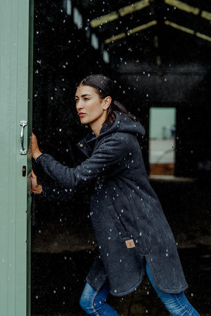 Wearing a Hard Yakka Womens Longline Zoodie with sherpa bonded fleece, a woman pushes open a green door in the rain, pausing partially outside as water droplets fall and a dark building rises in the background.