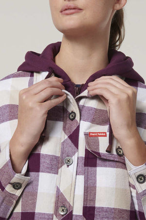 A person wears a purple hoodie under the Hard Yakka Women's Quilted Shacket, showcasing a checkered pattern in purple, beige, and white. They hold the buttons of the shacket, which features a red and white label on its pocket.