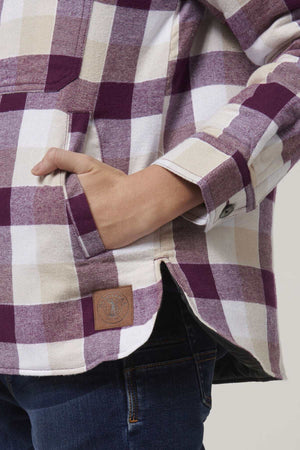 A person in the Hard Yakka Women's Quilted Shacket, purple and white plaid with quilted lining, a hand in the pocket, paired with dark jeans. The shacket features a small brown patch near the bottom hem.