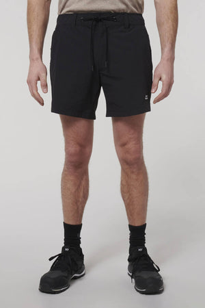 Facing forward, a person wears Hard Yakka X Short Short, black crew socks, and black sneakers. The upper body is not visible, and the background is plain and light-colored.