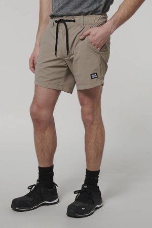 Someone stands against a gray background wearing a gray shirt, black sneakers, and black socks, paired with Hard Yakka X Short Shorts. The beige knee-length shorts are made of Cordura Nylon, featuring a drawstring and a small black label on the left side.