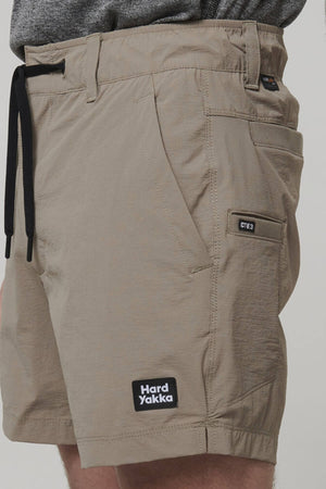 A model wears beige Hard Yakka X Short Shorts, made from durable Cordura Nylon with 4-way stretch and a small front logo patch. The shorts have multiple pockets and a black drawstring, set against a plain backdrop, with the model's torso partially visible.