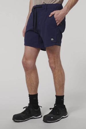 A person stands against a plain light background wearing Hard Yakka X Short Shorts in navy blue Cordura Nylon with a black drawstring, paired with black athletic shoes and socks, one hand in their pocket.