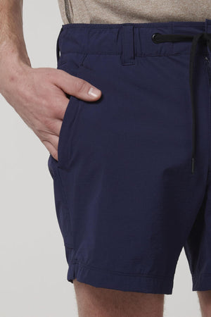 A person wears the Hard Yakka X Short Short in dark blue with a beige shirt, one hand in the shorts pocket, shown from waist to mid-thigh against a plain background.