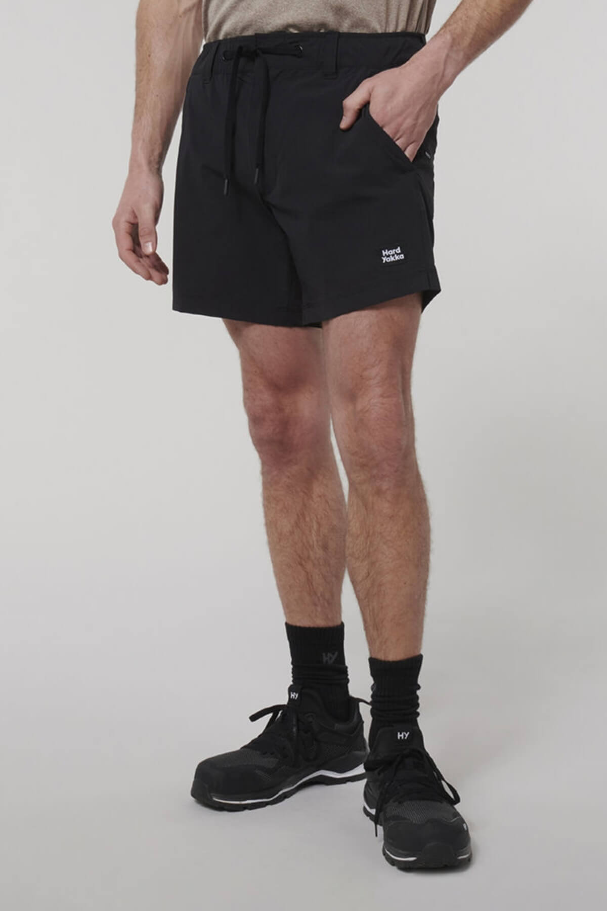 A person wears black Hard Yakka X Short Short athletic shorts with a drawstring waist, 4-way stretch fabric, and a small white logo on the left leg, paired with a beige shirt, black socks, and black sneakers against a plain background.
