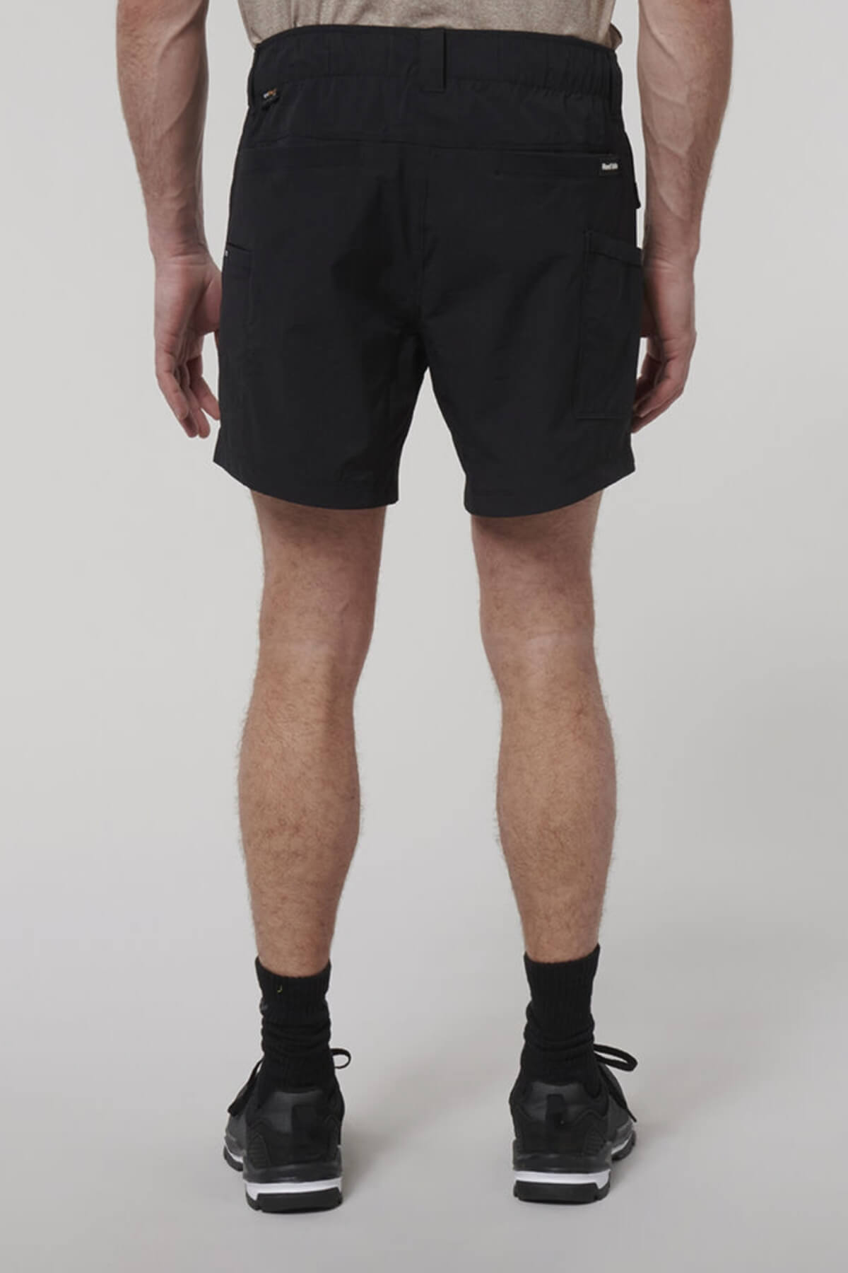 A person in black Hard Yakka X Short Shorts, black socks, and black sneakers stands with their back to the camera against a plain light background.