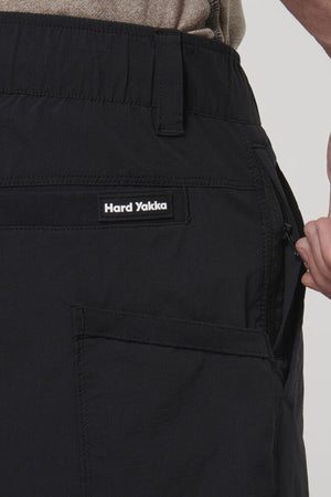 Close-up of a person wearing black Hard Yakka X Short Short pants with 4-way stretch Cordura Nylon, hand pulling the zipper on a side pocket. The Hard Yakka label is visible above the pocket.