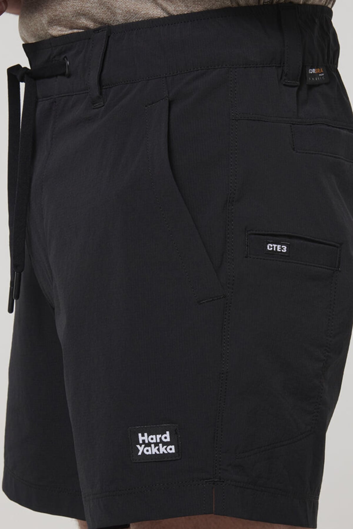 A close-up of a person wearing black Hard Yakka X Short Short with 4-way stretch, belted waist, front pockets, small CTE3 label, Cordura Nylon panels, and the Hard Yakka logo patch near the hem.