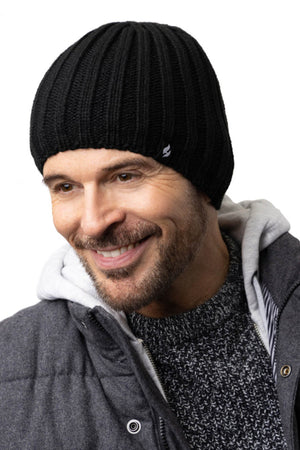 A man is smiling and wearing a black Heat Holders Halden Mens Thermal Beanie, a dark gray sweater, and a gray jacket over a hoodie. The background is plain and white.
