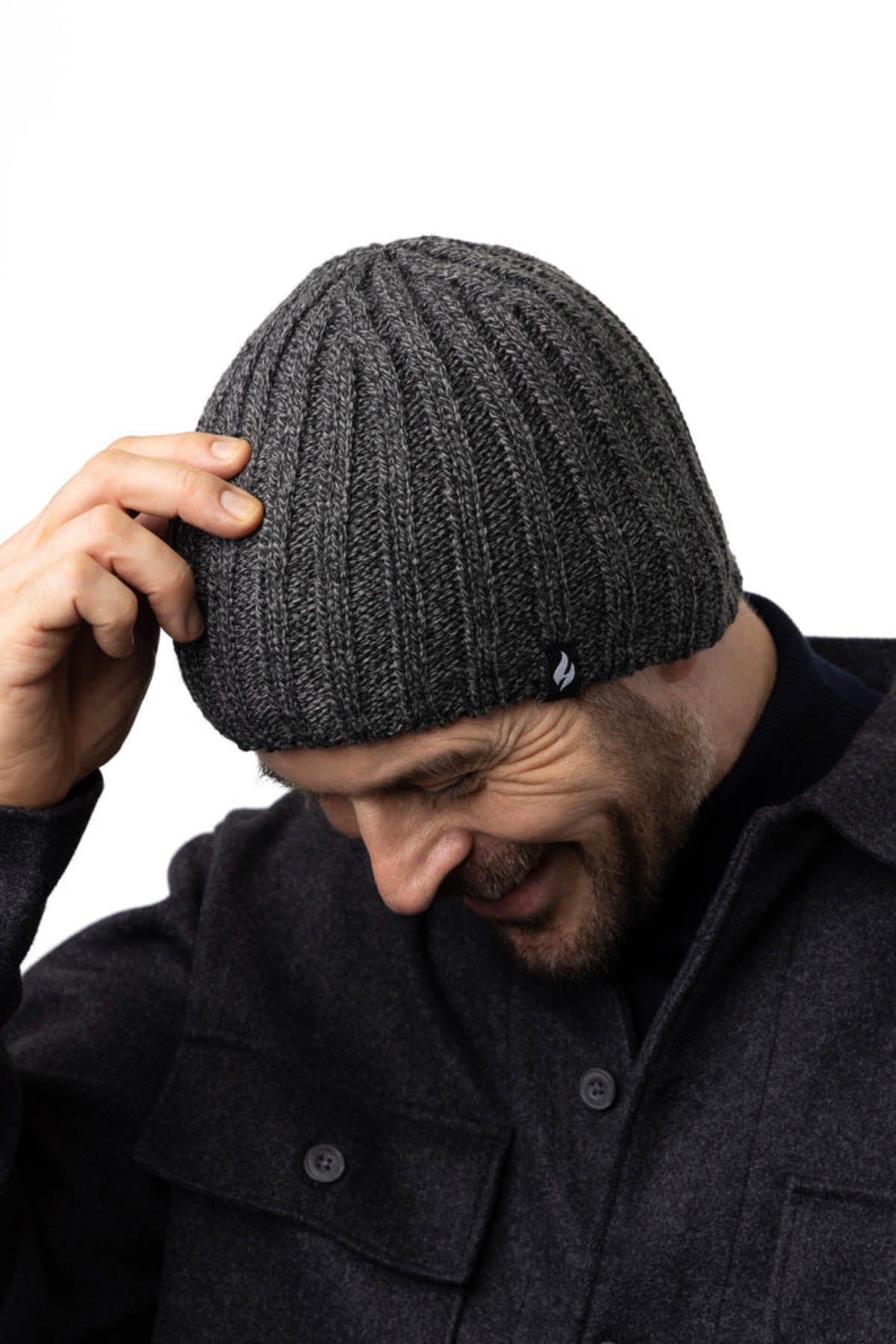 A man is smiling and looking down while adjusting a Heat Holders Halden Men's Thermal Beanie on his head. He is wearing a dark gray wool coat with large buttons, perfect for outdoor winter activities. The background is plain and white.