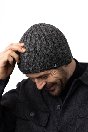 A man is smiling and looking down while adjusting a Heat Holders Halden Men's Thermal Beanie on his head. He is wearing a dark gray wool coat with large buttons, perfect for outdoor winter activities. The background is plain and white.