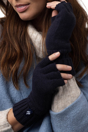 A woman wears a cozy blue sweater and light beige scarf, showcasing navy blue Heat Holders Ladies Fingerless Thermal Gloves designed for outdoor activities. Her long brown hair is down, and she is gently touching her left wrist with her right hand, emphasizing the HeatWeaver Thermal Lining's texture and fit.