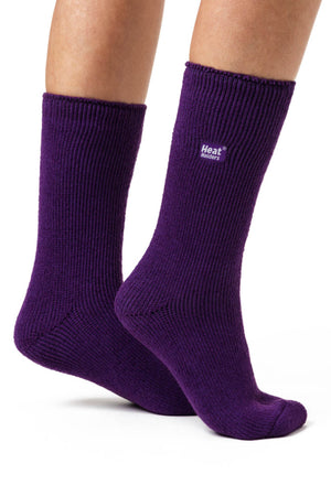 A pair of feet are shown wearing purple, ribbed Heat Holders Ladies Original Thermal Heat Socks. One foot is flat on the ground, and the other is slightly raised, displaying both the side and sole of the socks. The background is plain white. These socks boast an impressive TOG rating for maximum warmth.