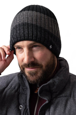 A man with a beard is wearing a black and gray knit Heat Holders Linden Block Stripe Mens Thermal Beanie and a dark gray jacket, showcasing the perfect winter outfit. He is holding the edge of his thermal beanie with one hand and looking slightly to the side. The background is white.