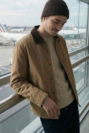 A man wearing a Hechter Patrick Canvas Jacket, beige sweater, and dark beanie stands by an airport window, smiling slightly as he looks down. Airplanes can be seen outside on the runway.