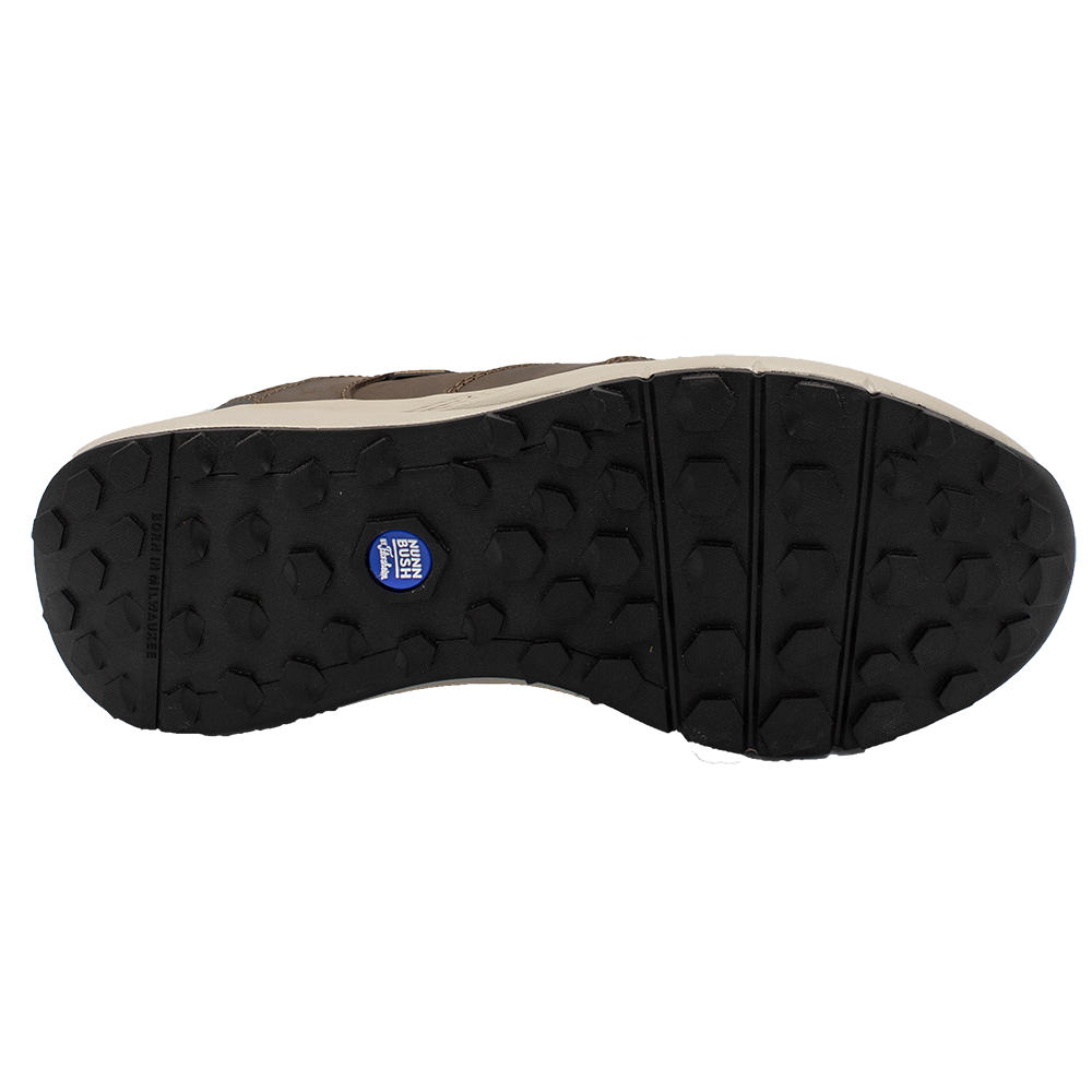 The image shows the sole of a Florsheim Excursion Slip On shoe with a rugged, textured pattern for shock absorption, featuring hexagonal grips and a small blue logo in the center. The sole is primarily black with brown detailing along the edges.