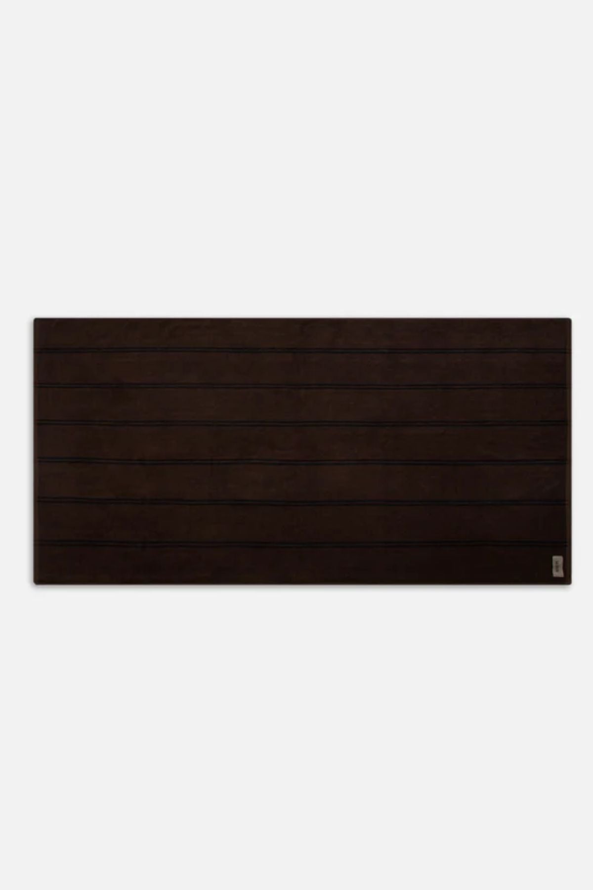 A rectangular, dark brown wooden surface with horizontal grooves and a small metal label in the bottom right corner sits against a plain white background, evoking the look of Industrie The Comporta Beach Towelβs cotton terry texture.