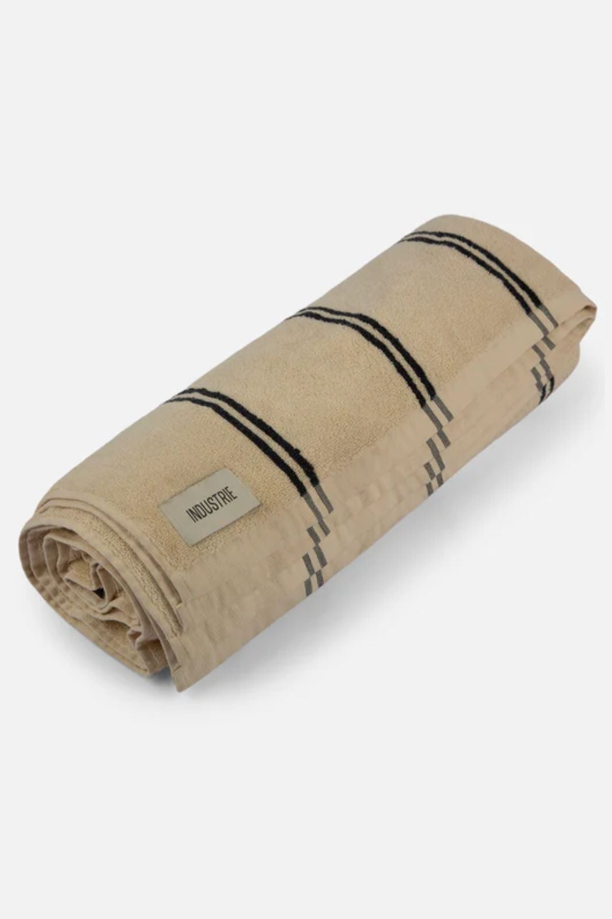 The Industrie The Comporta Beach Towel is a beige, soft cotton terry cloth towel with thin black stripes and a visible "INDUSTRIE" label, neatly rolled and set against a light grey background.