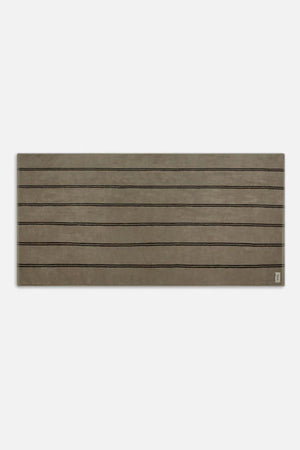 The Industrie Comporta Beach Towel is rectangular, brown, and features thin black horizontal lines inspired by halo stripes with a small logo in the bottom right corner.