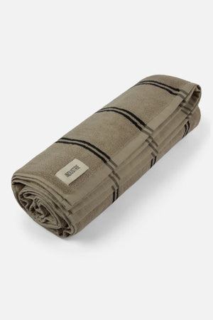 The Industrie The Comporta Beach Towel is a neatly rolled beige cotton terry cloth towel with black plaid stripes and a white "MidoriTec" label on one end, displayed on a plain light background.