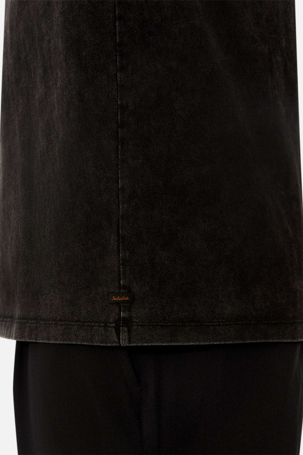 Close-up of the lower half of the Industrie The Del Sur Acid Wash Tee in faded black with an oversized fit over black pants, featuring a small brown embroidered label near the hem. Background is plain white.