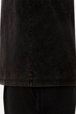 Close-up of the lower half of the Industrie The Del Sur Acid Wash Tee in faded black with an oversized fit over black pants, featuring a small brown embroidered label near the hem. Background is plain white.
