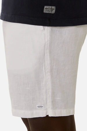 A person wears white linen-cotton shorts and a black Industrie The Del Sur Sleeveless Tee; only the waist to mid-thigh area is shown, highlighting small "Industrie" brand labels on both the shorts and the tee.