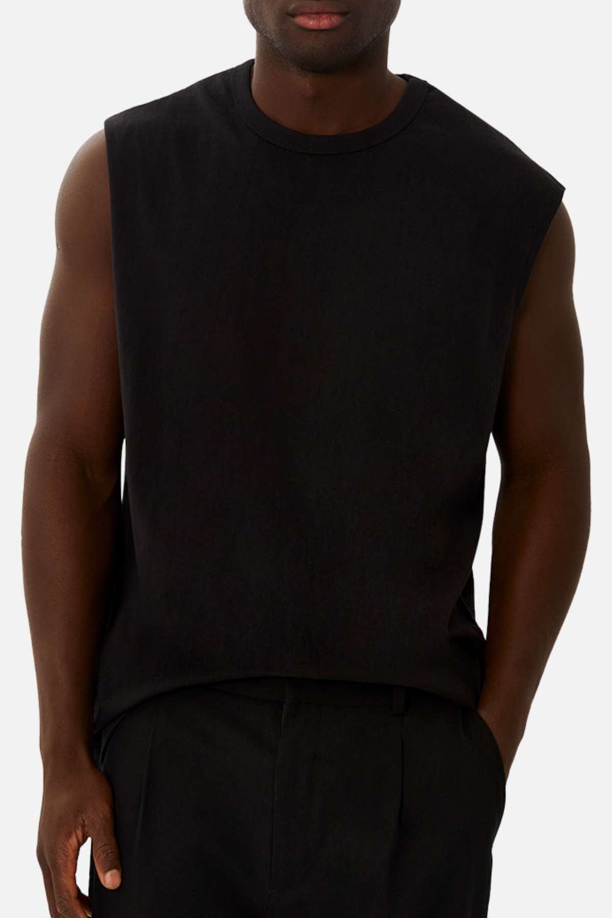 A man wearing the Industrie The Del Sur Sleeveless Tee in black 100% cotton and black pants stands with one hand partially in his pocket against a plain light background.